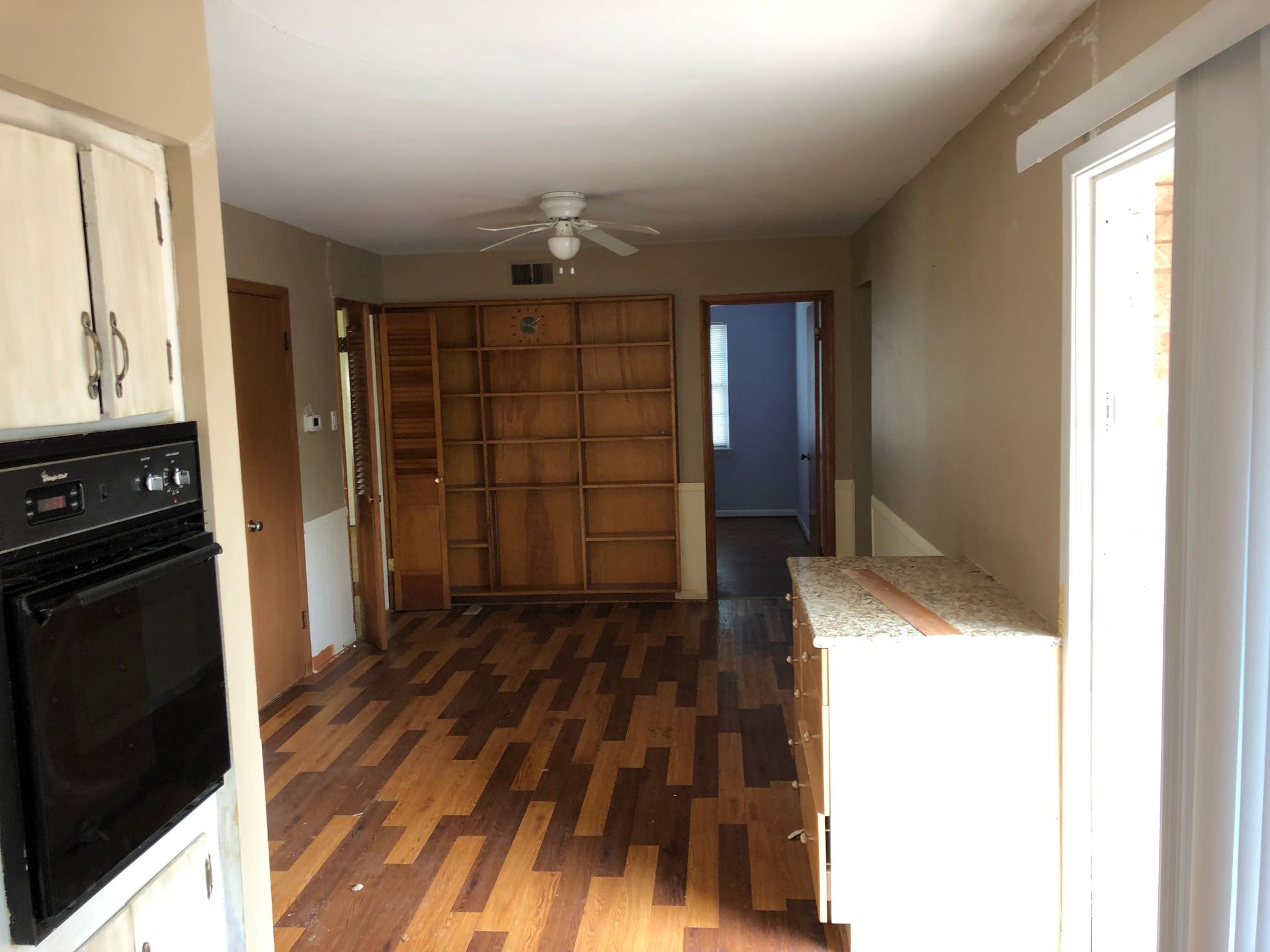 An interior view of a room with hardwood floors, a built-in wooden bookshelf, a black wall oven, and a sliding glass door.