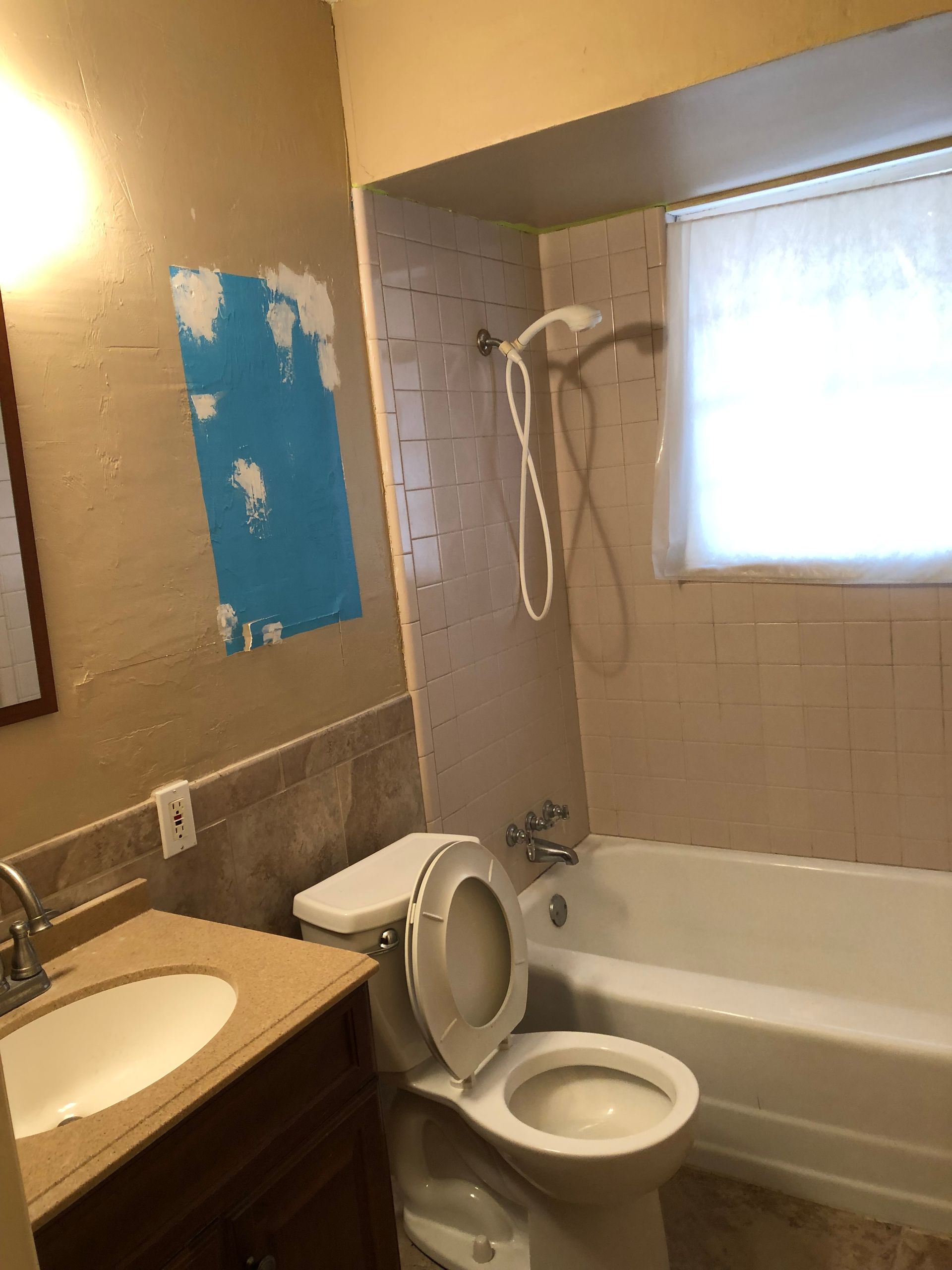 A bathroom with a vanity, toilet, and bathtub with a tiled shower wall, featuring a patch of blue wall repair material.