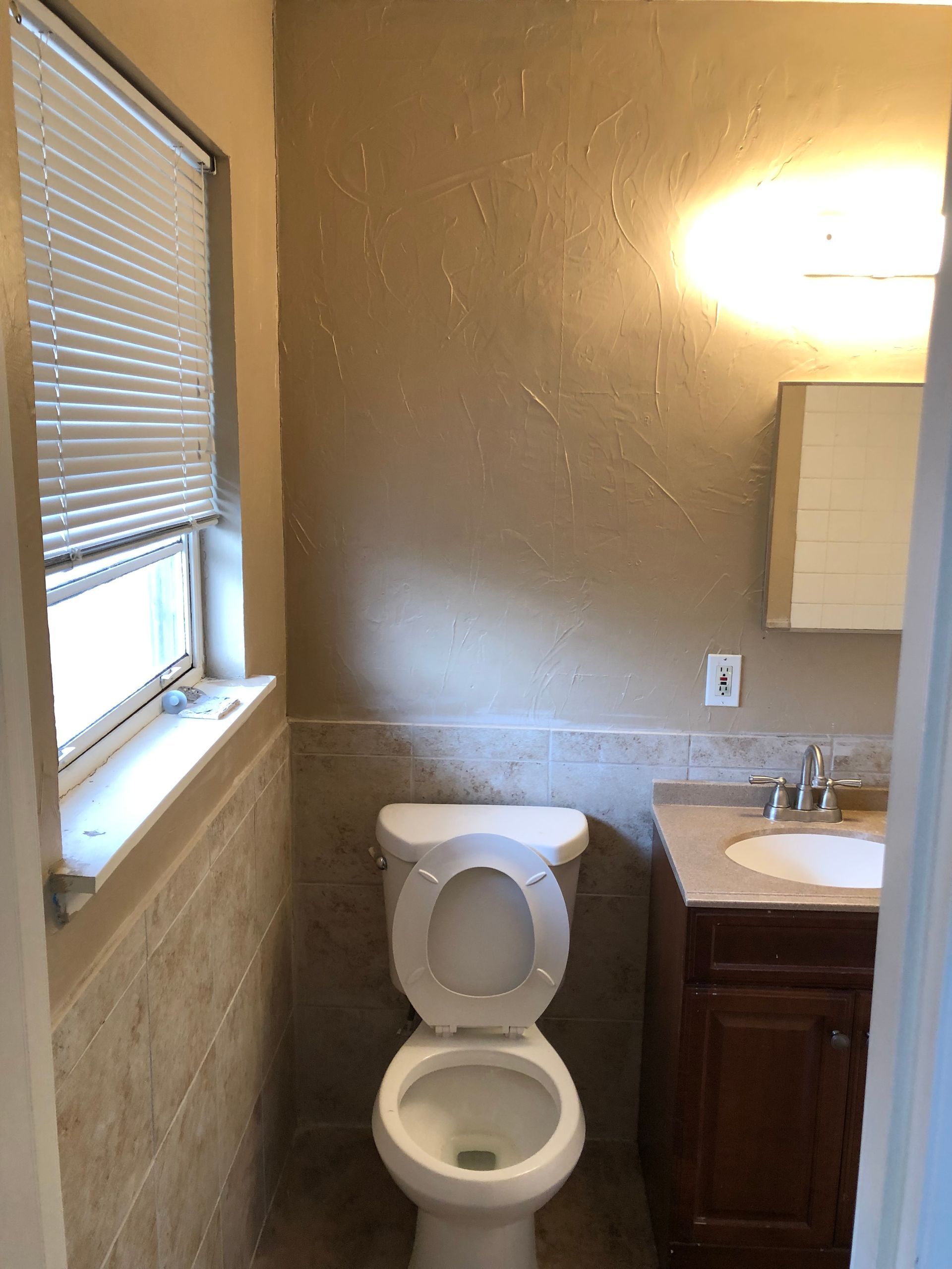 A small bathroom featuring a toilet, a vanity with a sink, beige textured walls, tiled lower walls, and a window with blinds.