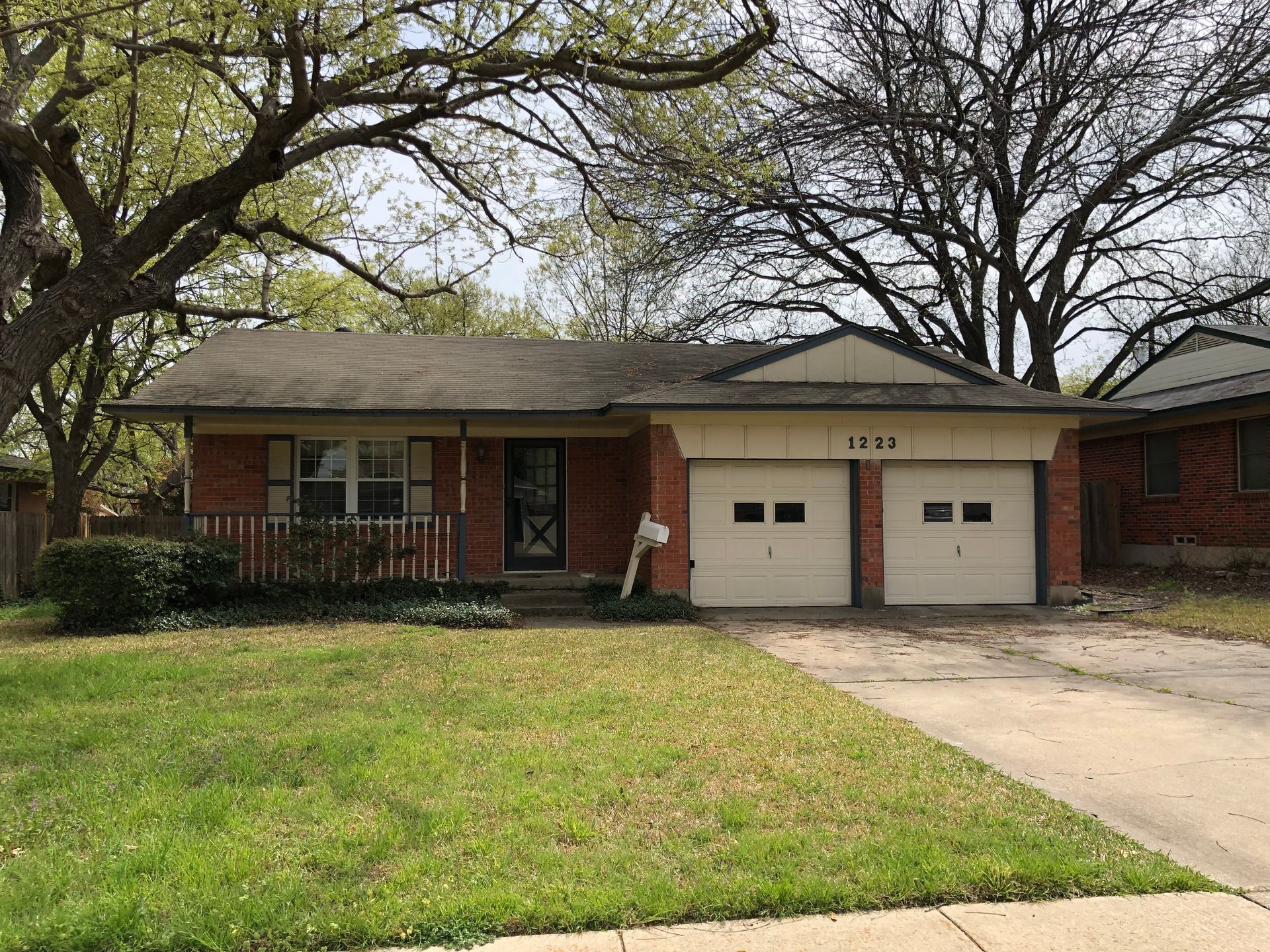 A single-story brick house with a two-car garage, white trim, and a small front porch, surrounded by trees and a lawn.