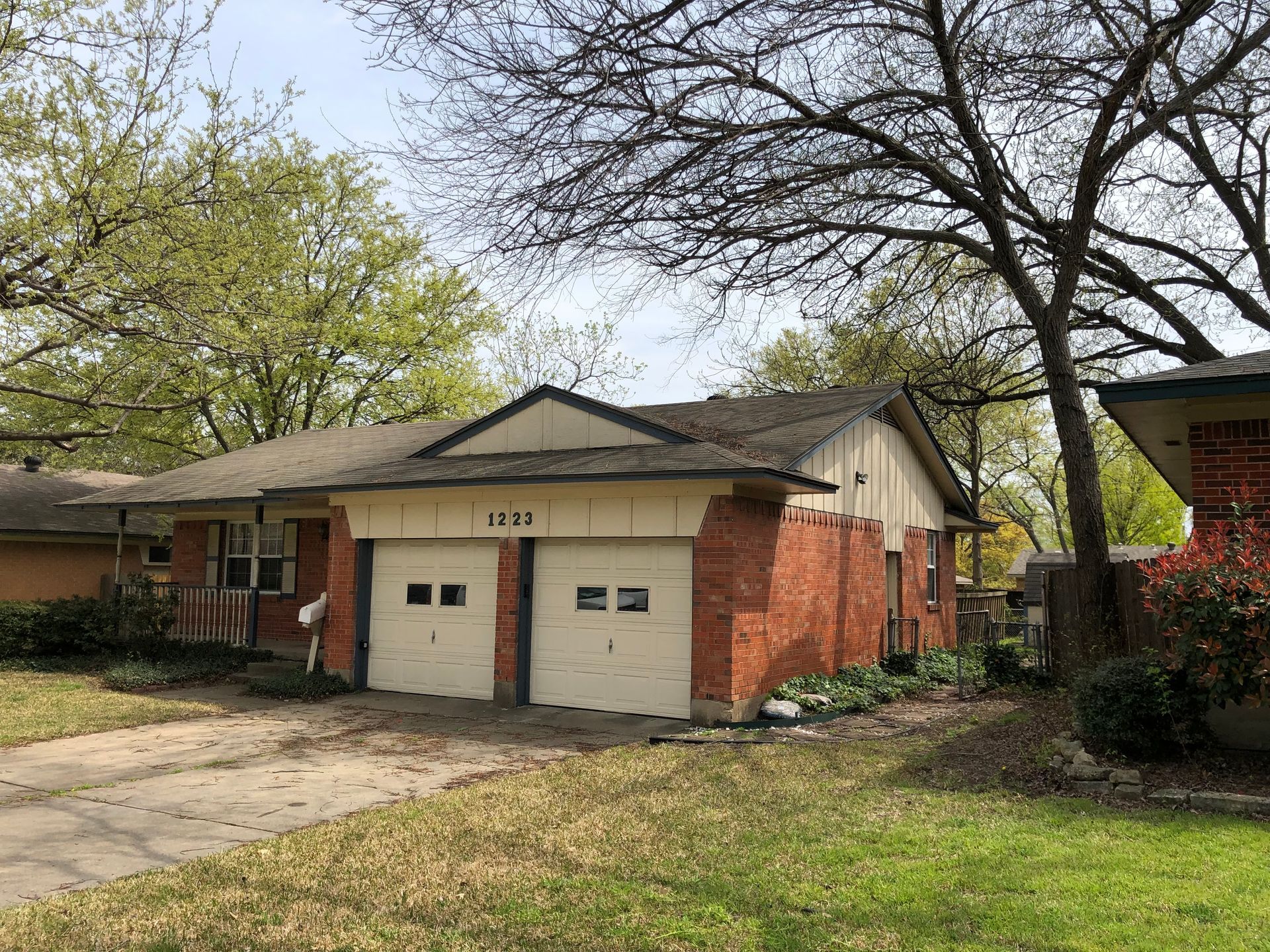 A single-story red brick house with a two-car garage, beige trim, and a yard with mature trees.