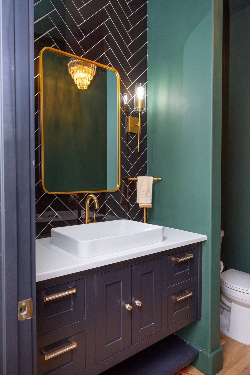 A small bathroom with a white sink on a dark blue vanity, a gold-framed mirror, and a black herringbone-tiled accent wall.