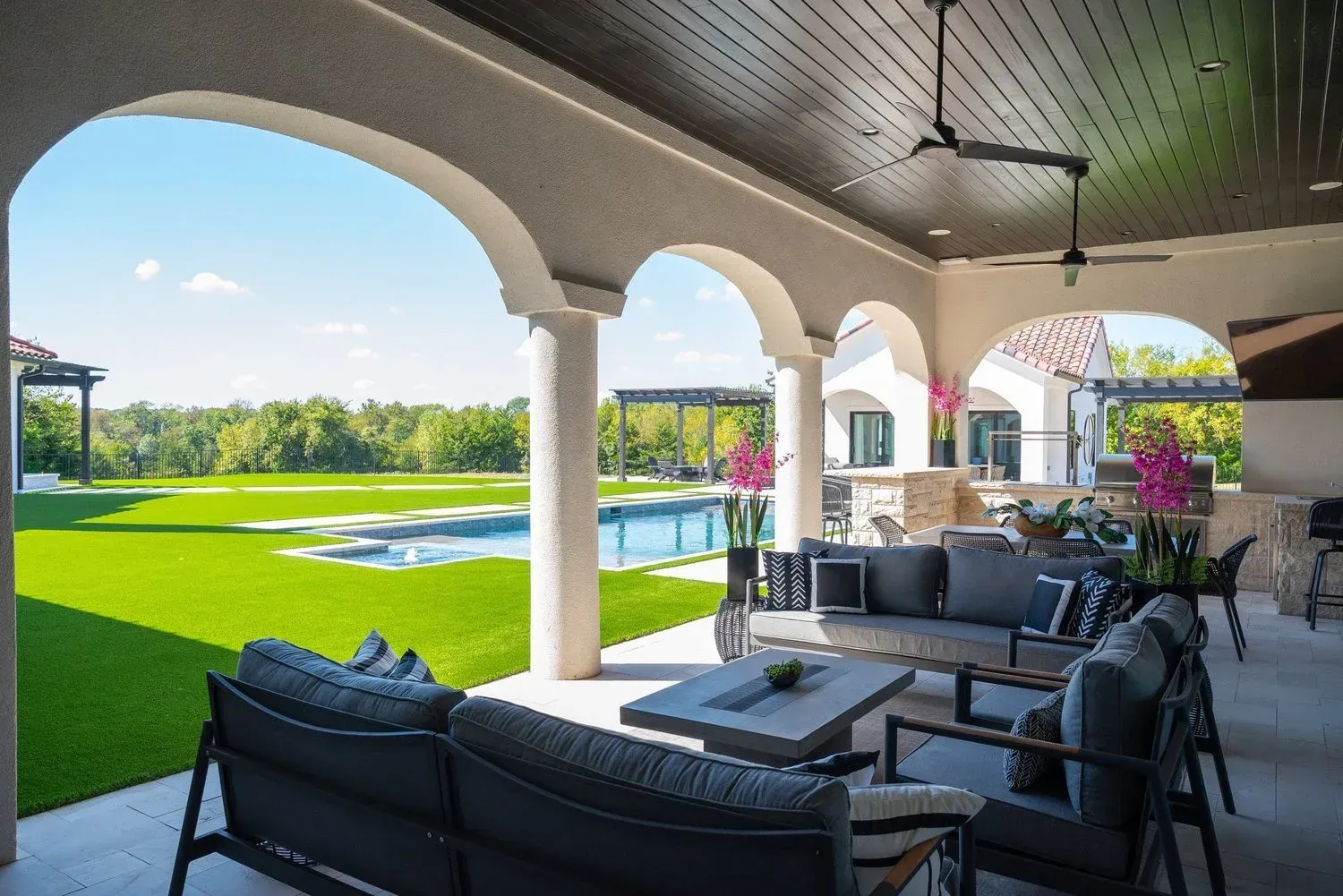 A covered outdoor patio featuring grey lounge furniture, a pool, and a grassy yard under a bright, sunny sky.