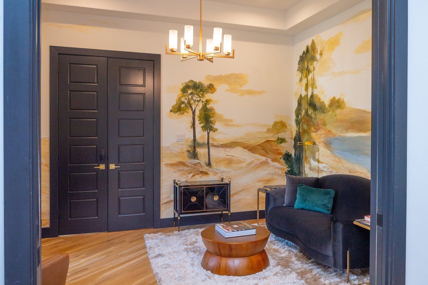 A lounge with a dark double door, a couch with a teal pillow, a round wooden table, and a mural of a landscape.