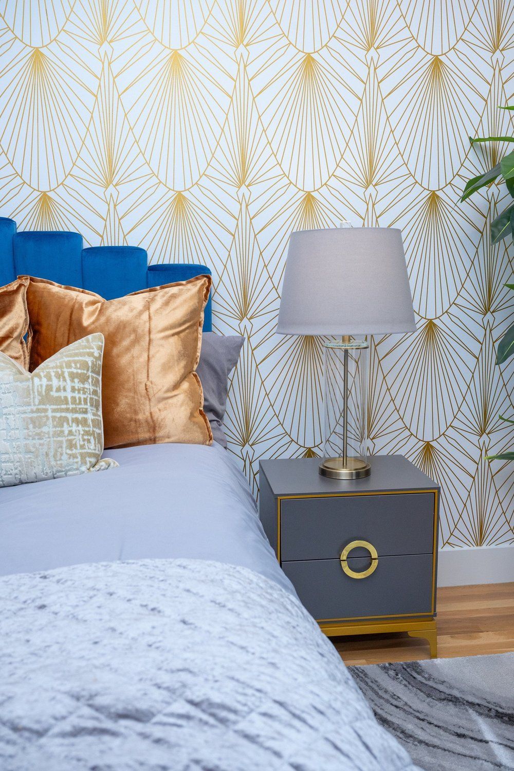 A blue velvet bed with gold pillows next to a grey nightstand and table lamp against gold and white patterned wallpaper.