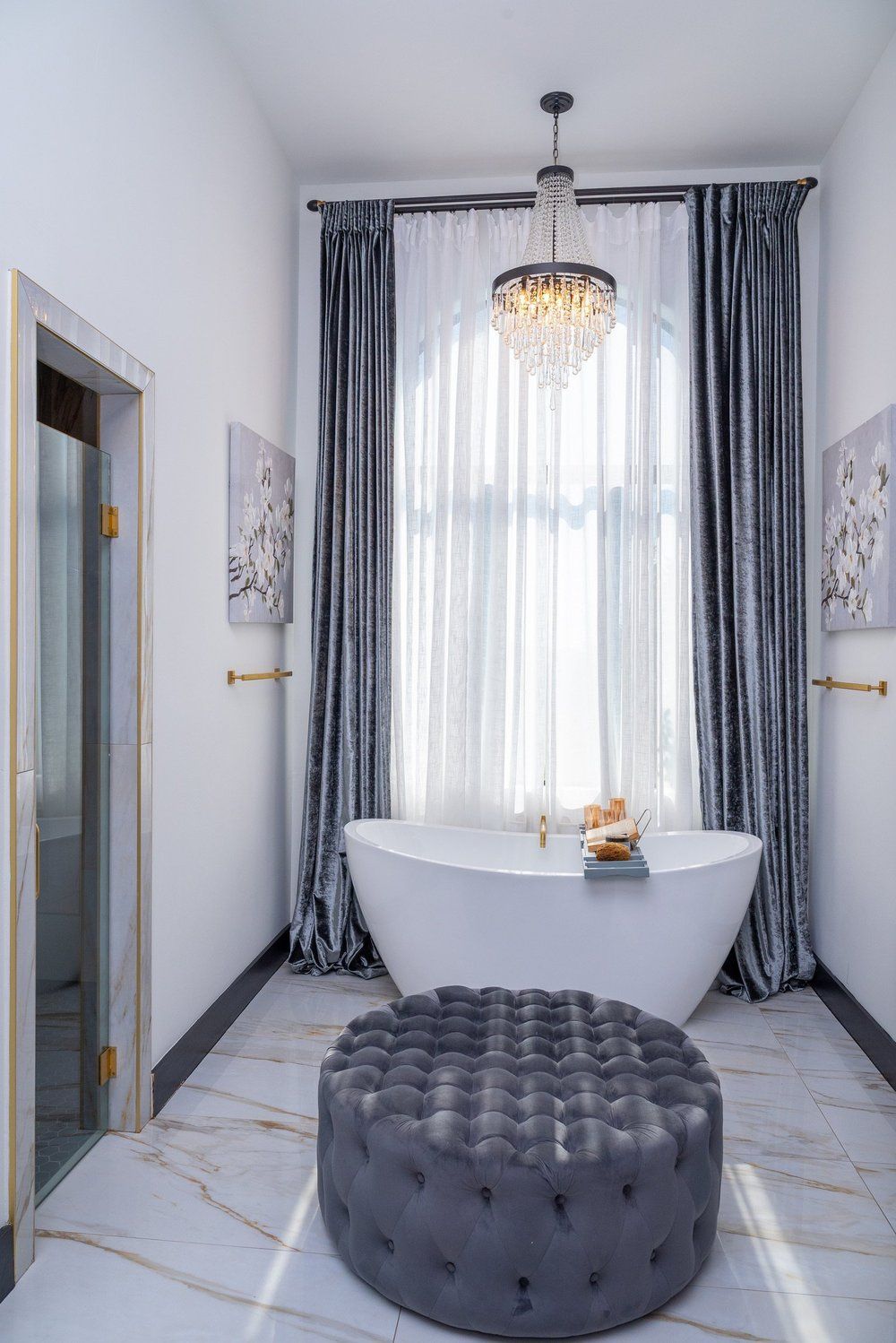 A bathroom with a freestanding white tub, a dark gray tufted ottoman, a crystal chandelier, and floor-to-ceiling curtains.