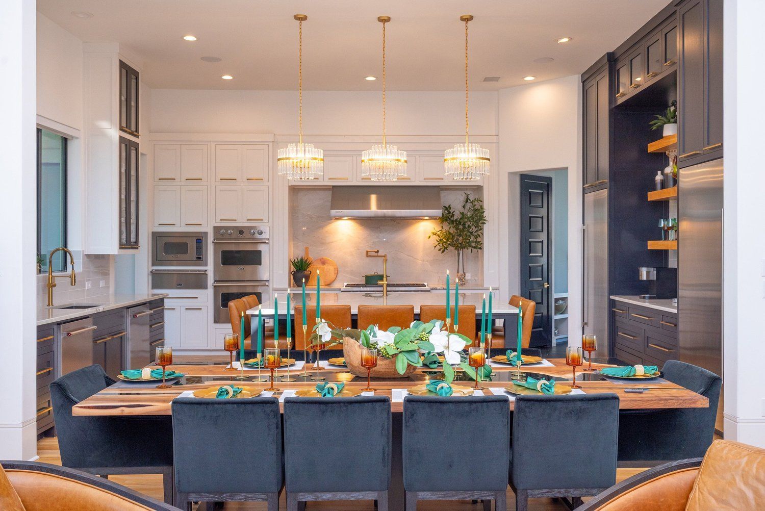 A modern kitchen with white cabinetry, a wooden dining table, dark blue chairs, and three gold pendant lights.