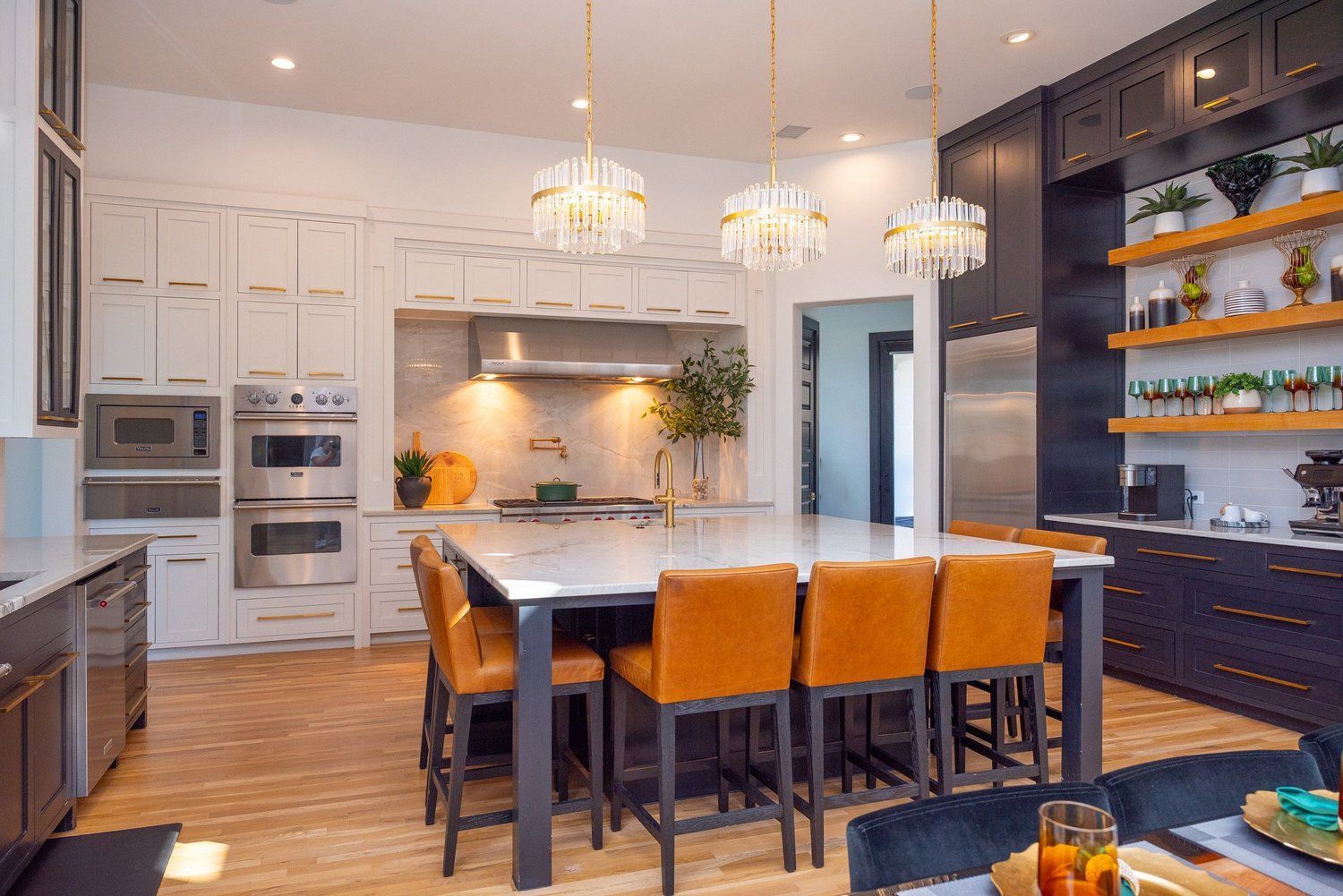 A modern kitchen featuring white cabinets, a large central island with tan stools, gold pendant lights, and open shelving.