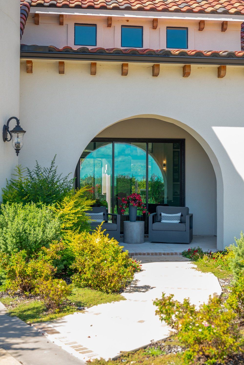 A white arched entryway with two chairs and a path leading through a garden towards a building with a terracotta roof.