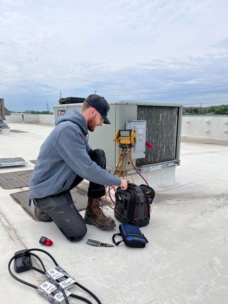 A man is kneeling down on a roof working on an air conditioner.