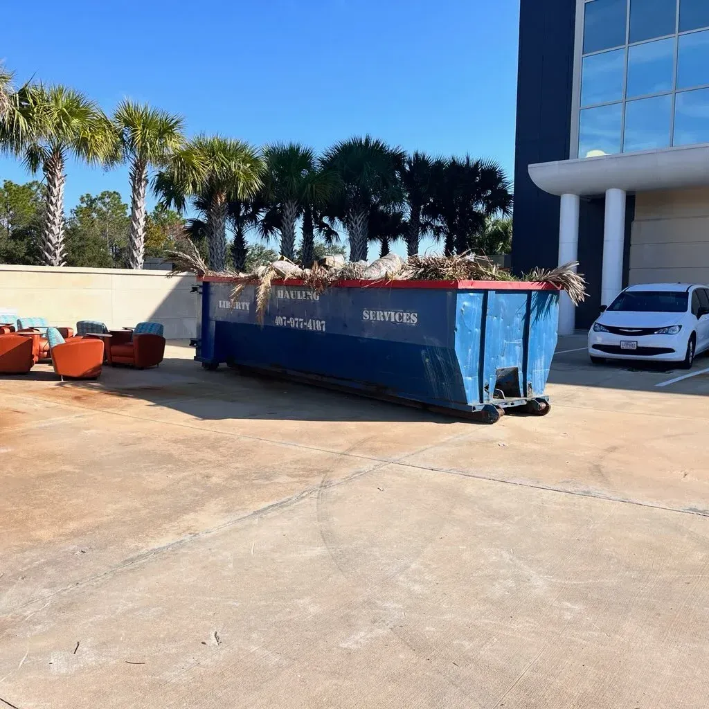 Blue dumpster beside a modern building with orange bins and a white car in bright sunlight