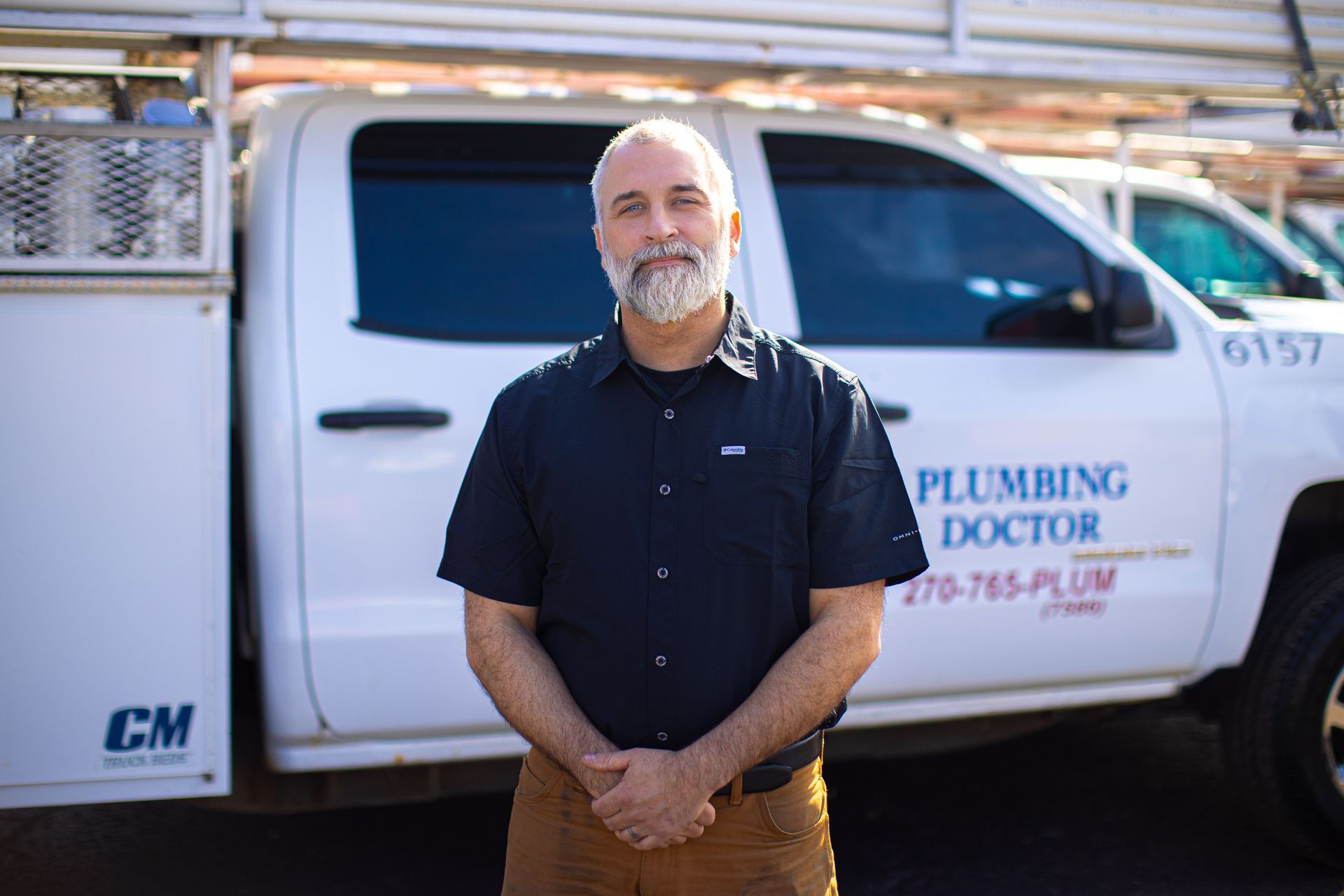 Man in front of a white plumbing truck, wearing a dark shirt and tan pants. Truck has 