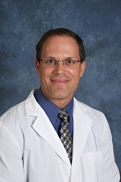 A man in a white lab coat and glasses smiles, wearing a tie and blue shirt.