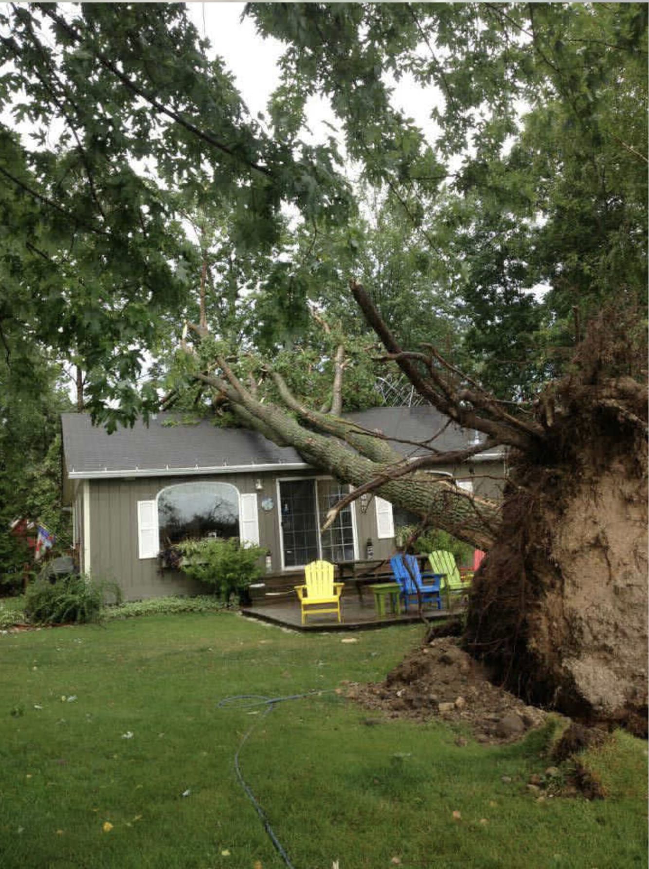 Uprooted tree fallen beside a small house, with a damaged yard and patio furniture visible.