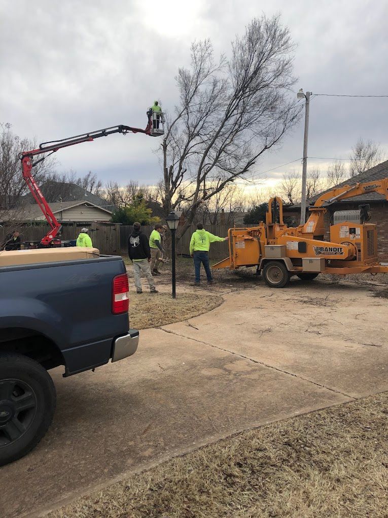 Workers in neon vests trimming a tree with a cherry picker beside a truck and wood chipper in a yard