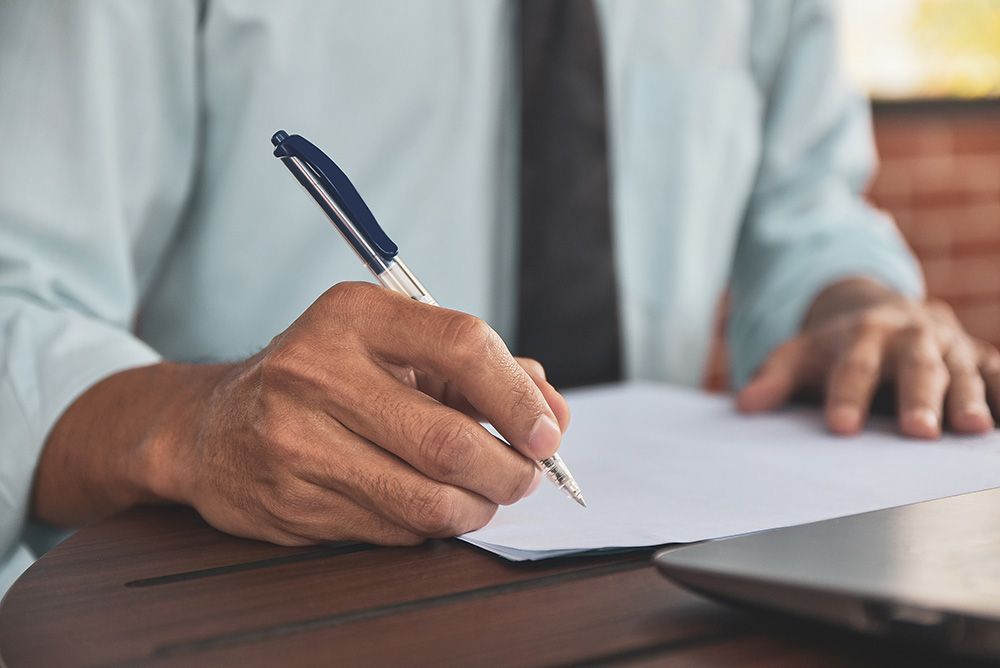 Person in light blue shirt and tie writing with a pen on white paper at a table.