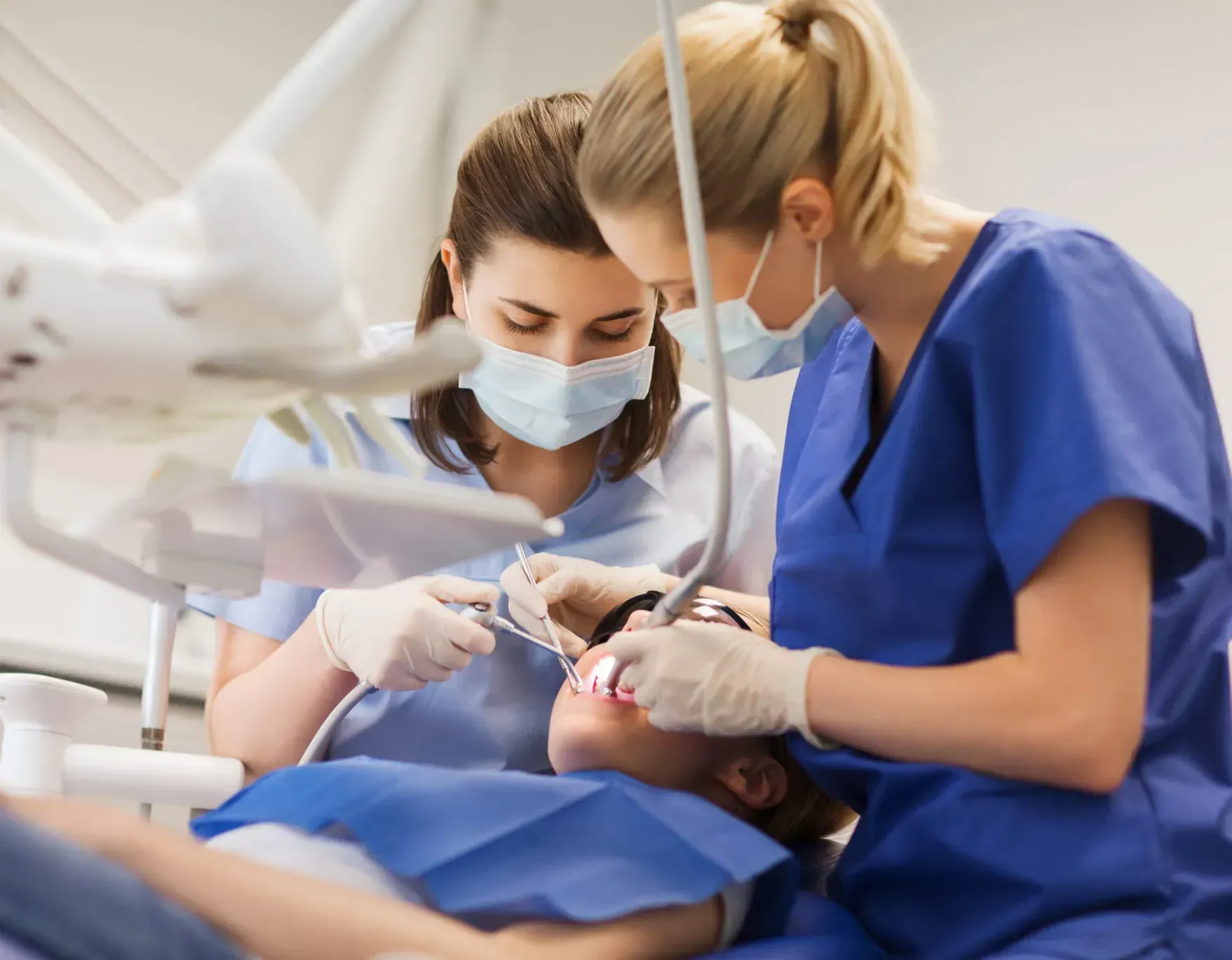 Dentist and assistant in blue scrubs treating a patient in a dental clinic, both wearing masks and gloves