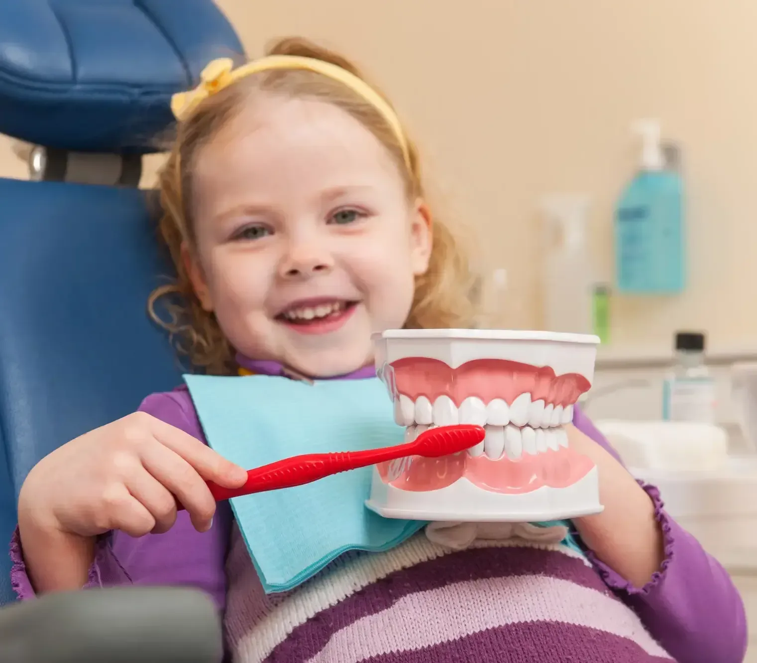 Child smiling in a dentist chair, brushing teeth on a dental model with a red toothbrush