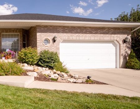 Suburban brick house with white garage door, driveway, and landscaped front yard under a blue sky