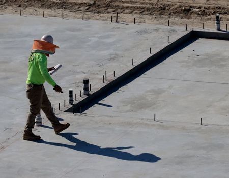 Worker in green shirt and hard hat walking across a concrete slab at a construction site.