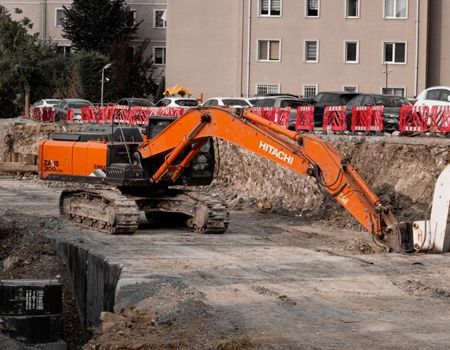 Orange excavator digging at a construction site beside a building and red barricades
