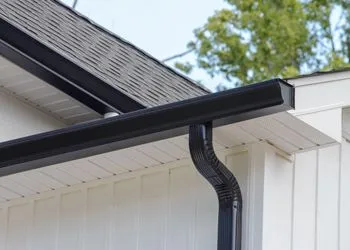 Black gutters on a white house, with a dark roof and a clear sky in the background.