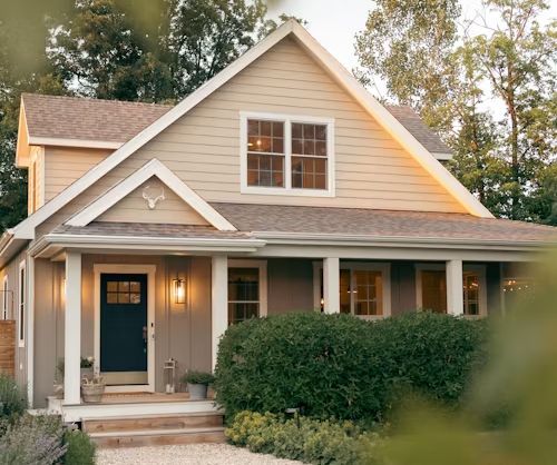 Charming beige house with blue front door, white trim, and a covered porch surrounded by shrubs and trees