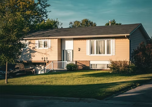 Single-story beige house with a front lawn and large tree in warm evening light
