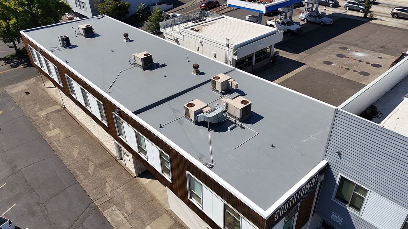 Aerial view of a long, flat building roof with HVAC units. The roof is gray and has several vents.