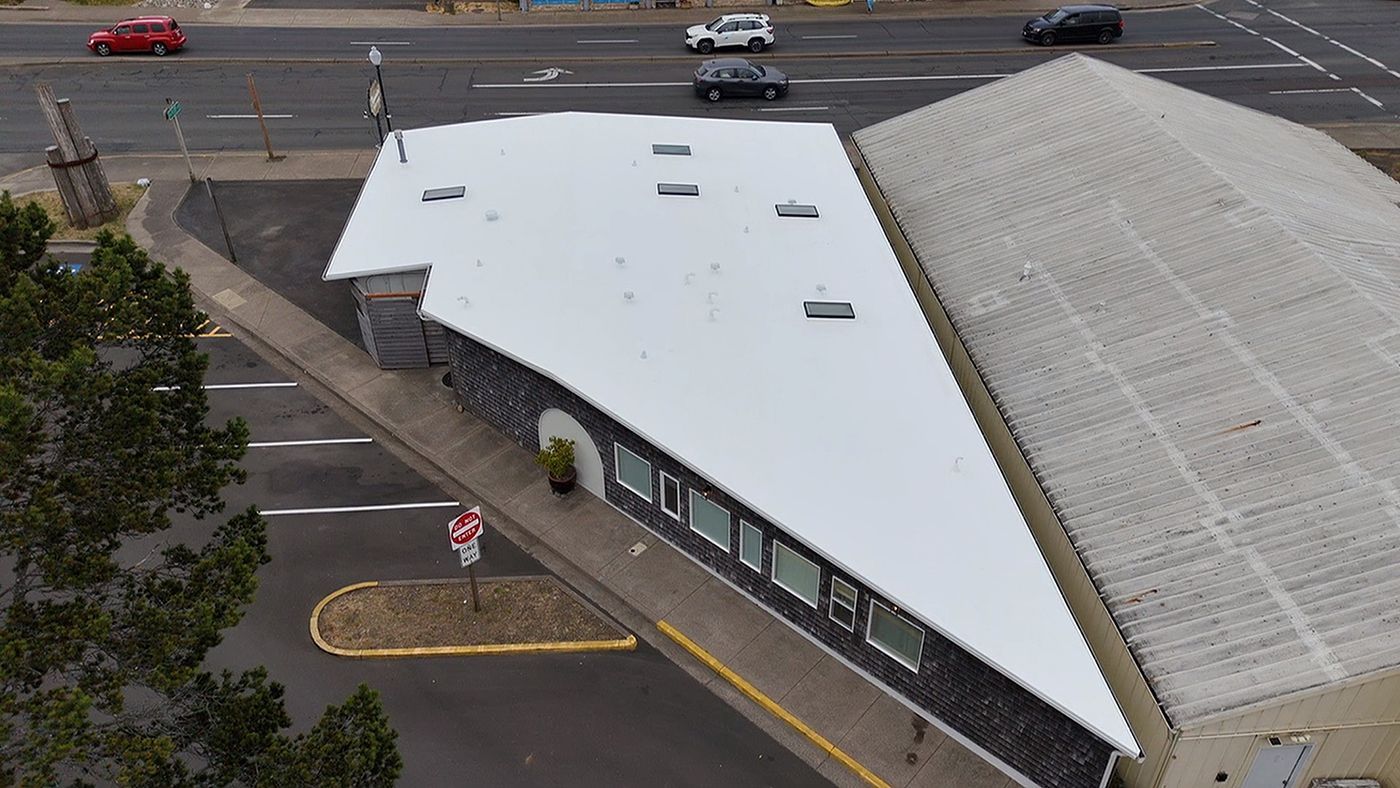 Aerial view of a building with a white roof, next to a building with a gray roof; cars drive on a nearby street.
