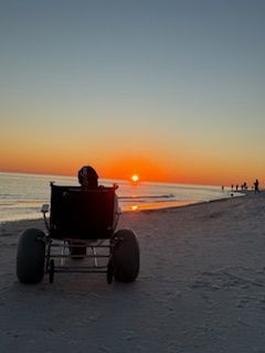 Our daughter in her beach wheelchair on a beach at sunset, facing the ocean with orange light on the horizon.