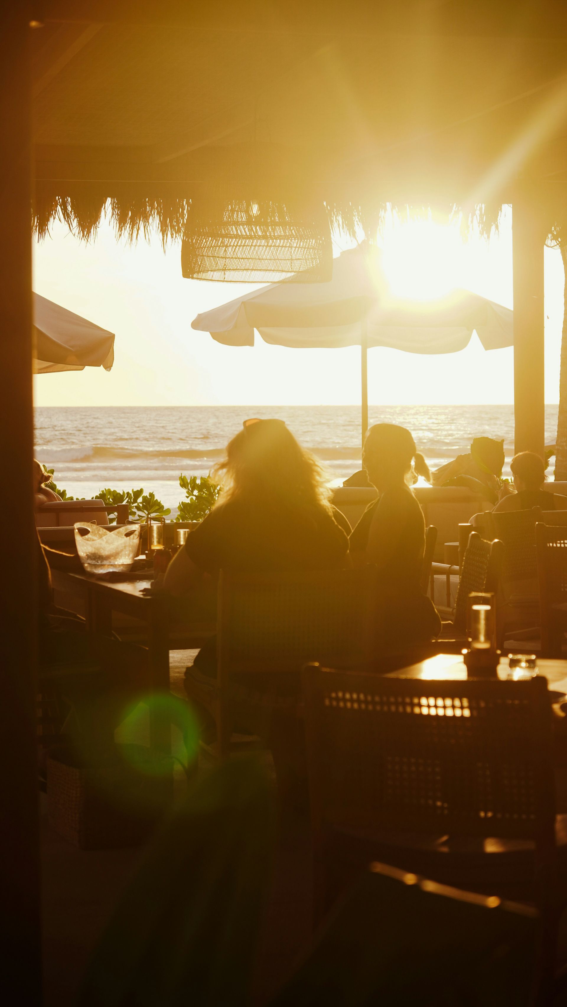 Sunlit beach café interior with silhouetted diners, umbrellas, and ocean view at sunset