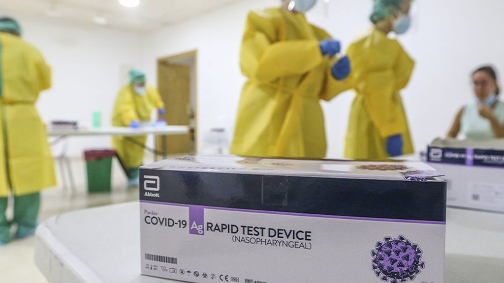 Una caja de kits de prueba rápida de COVID-19 se encuentra sobre una mesa, con trabajadores de la salud con equipo de protección en un área de prueba.