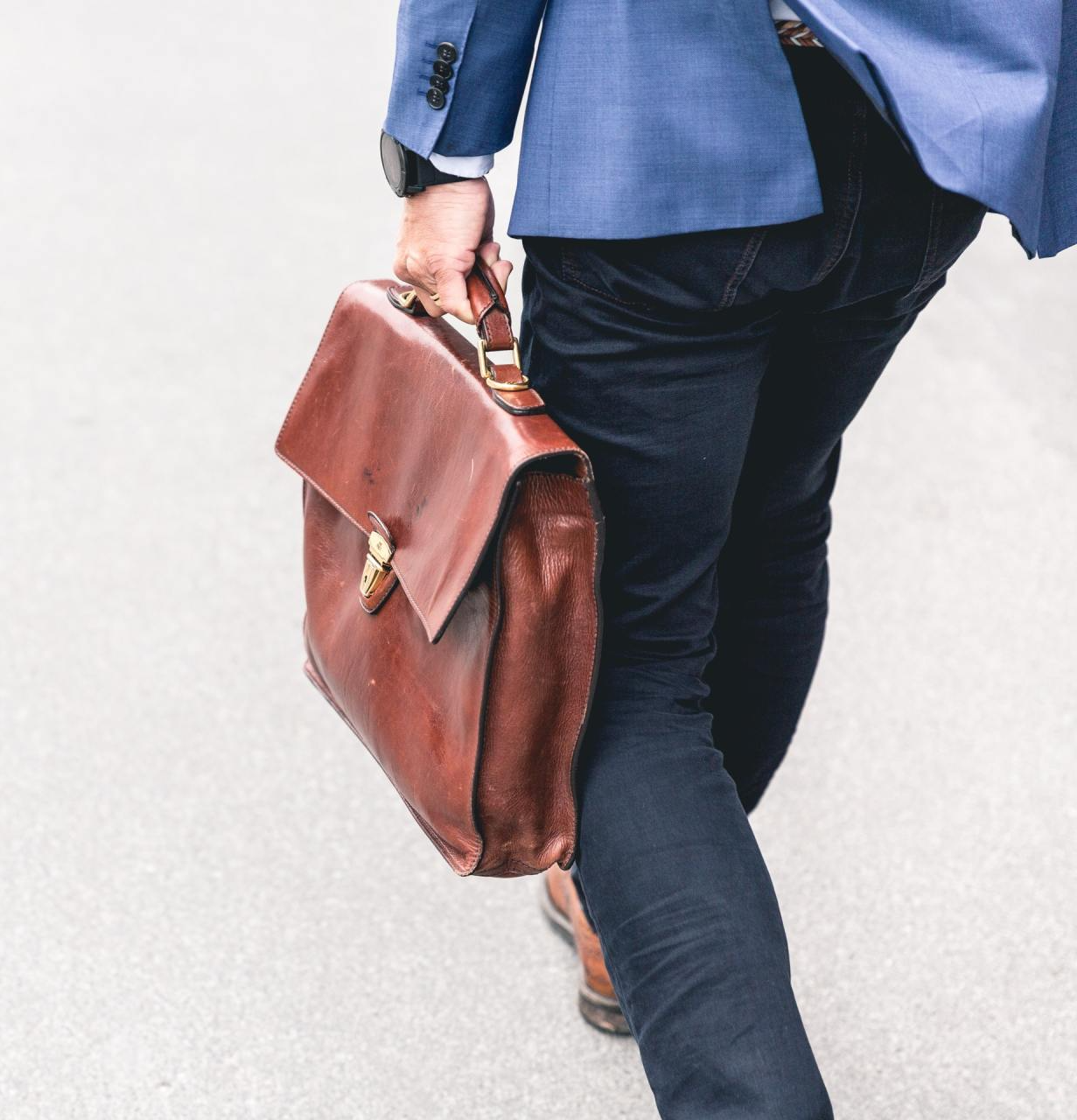 Persona caminando, sosteniendo un maletín de cuero marrón, vistiendo un blazer azul y jeans oscuros.