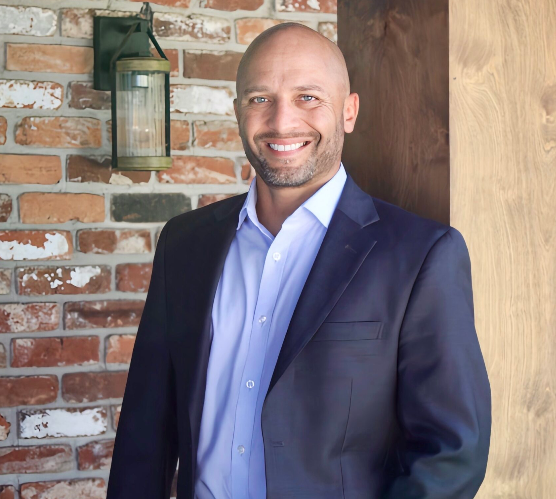 A man in a suit and blue shirt is standing in front of a brick wall.
