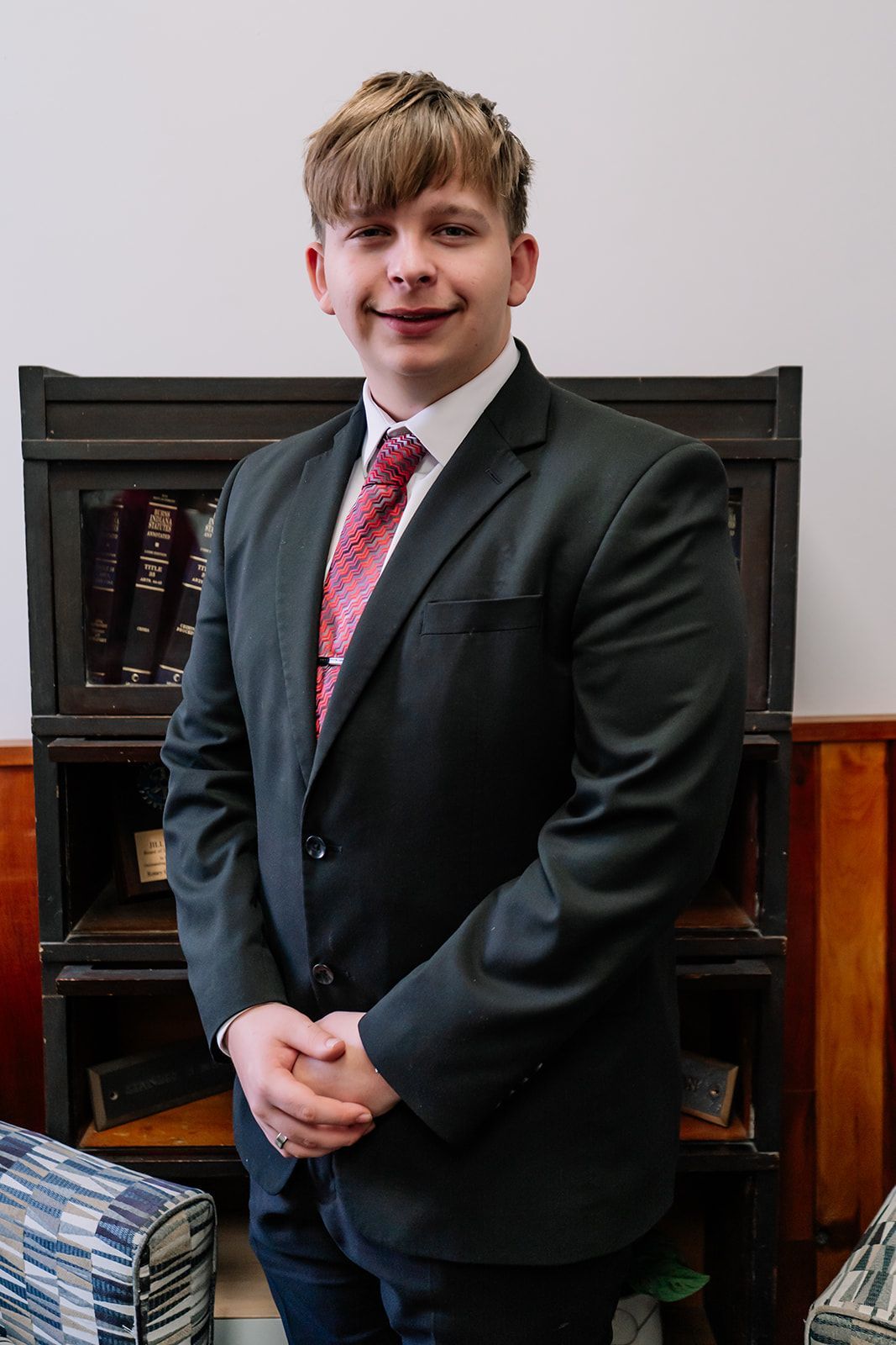 A young man in a suit and tie is standing in front of a bookshelf.