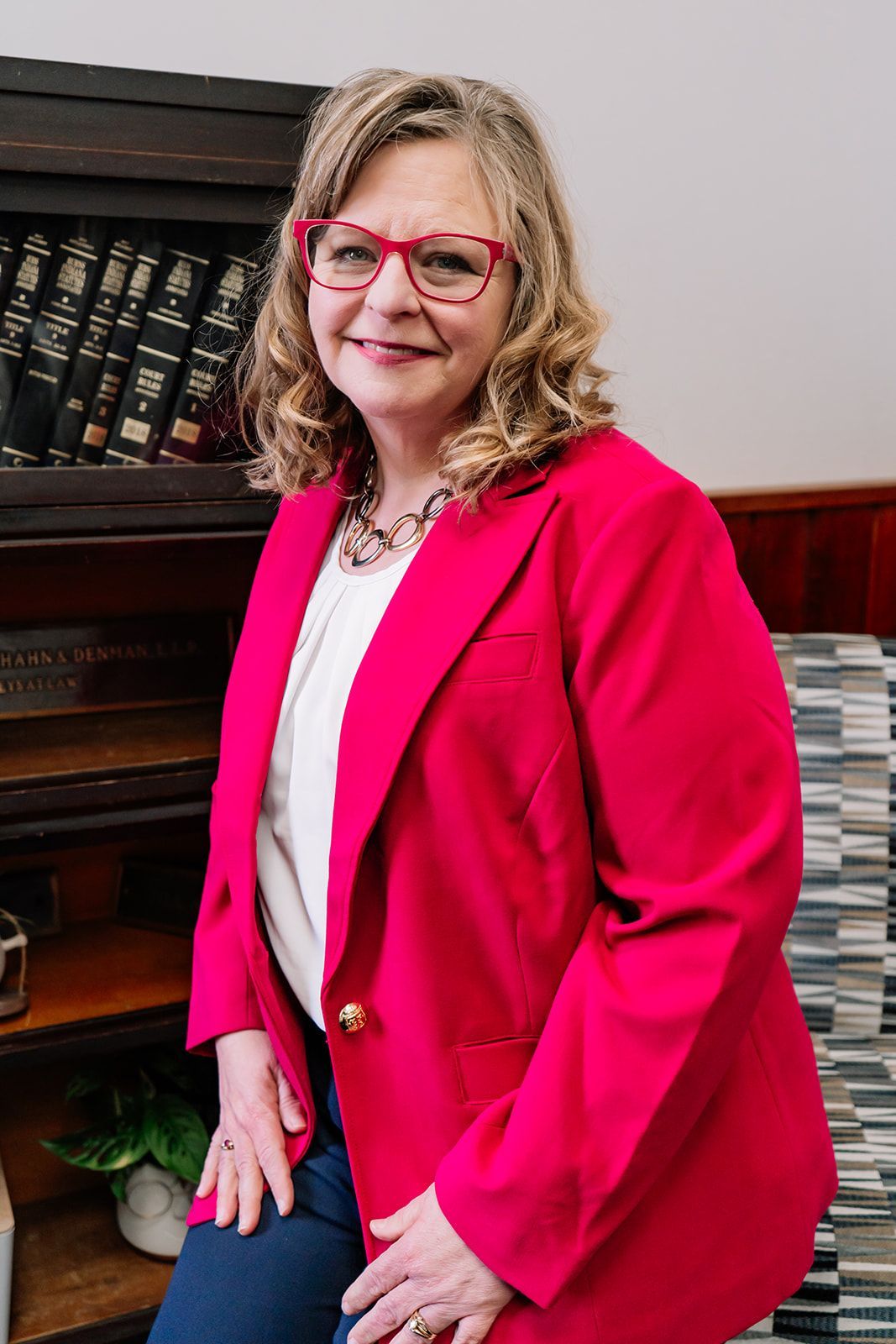 A woman in a pink jacket and glasses is sitting on a chair in front of a bookshelf.