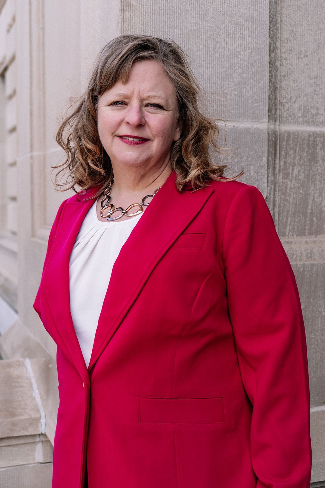 A woman in a red jacket and white shirt is standing in front of a building.