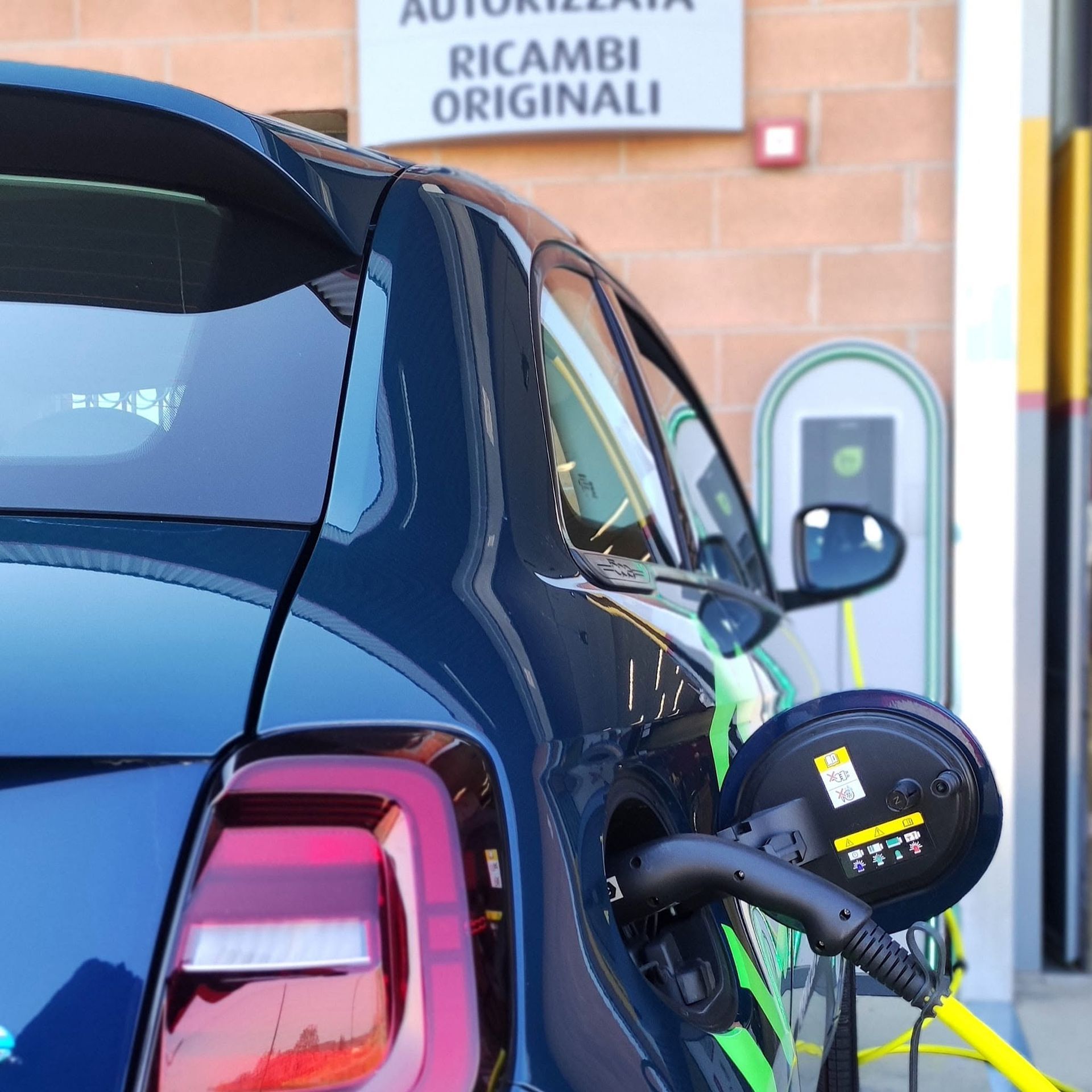 Auto elettrica blu in carica presso una stazione accanto a un muro di mattoni e un cartello con la scritta