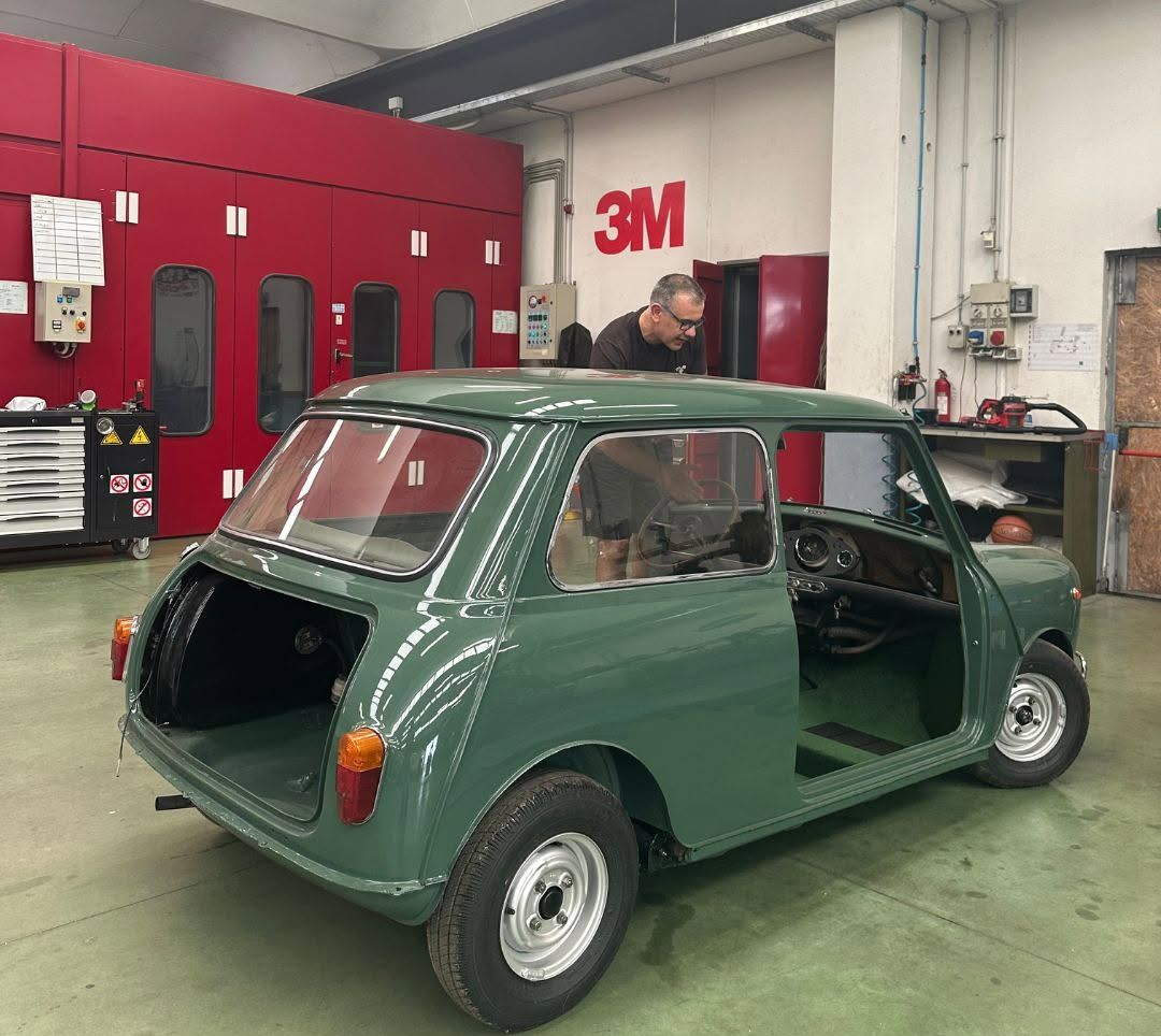 Una classica Mini Cooper verde in officina, con il portellone posteriore aperto e un meccanico al lavoro nelle vicinanze.
