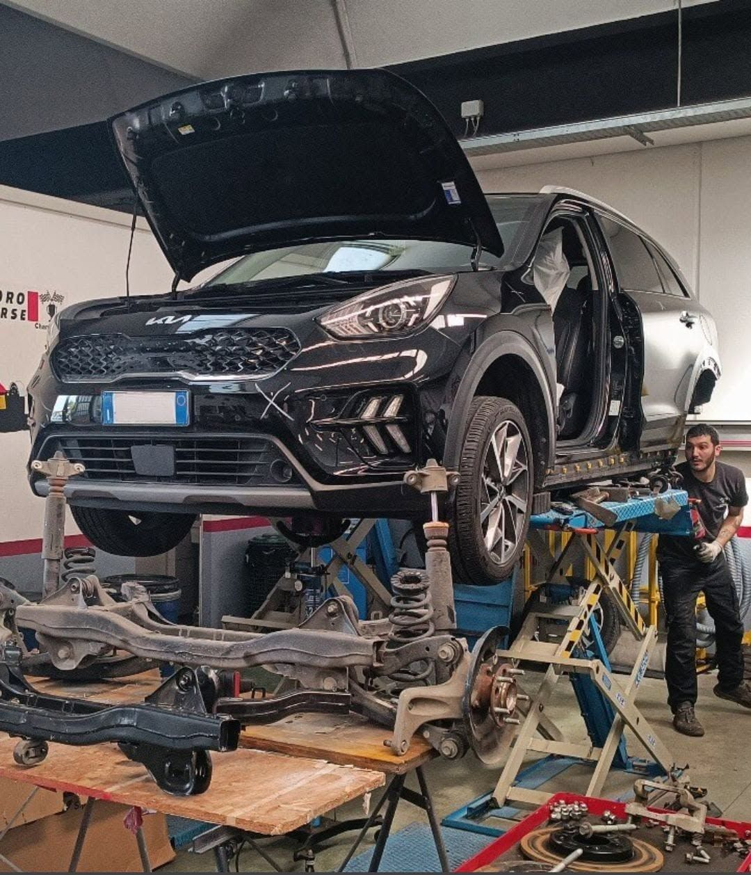 SUV nero con cofano aperto su un ponte sollevatore in un'officina meccanica affollata, con meccanici al lavoro nelle vicinanze.