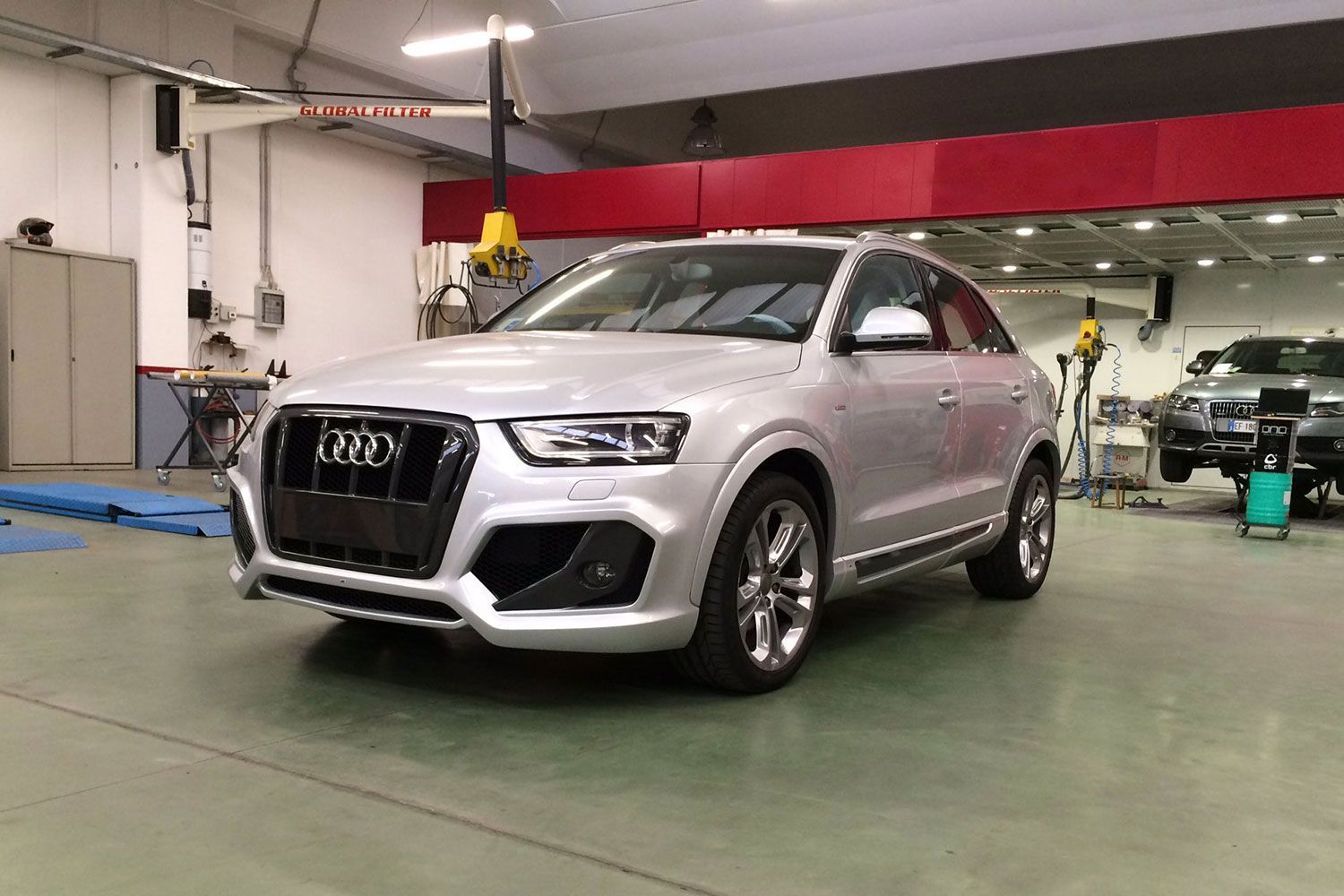 Un SUV Audi color argento in un'officina meccanica, parcheggiato su un pavimento verde sotto travi rosse.