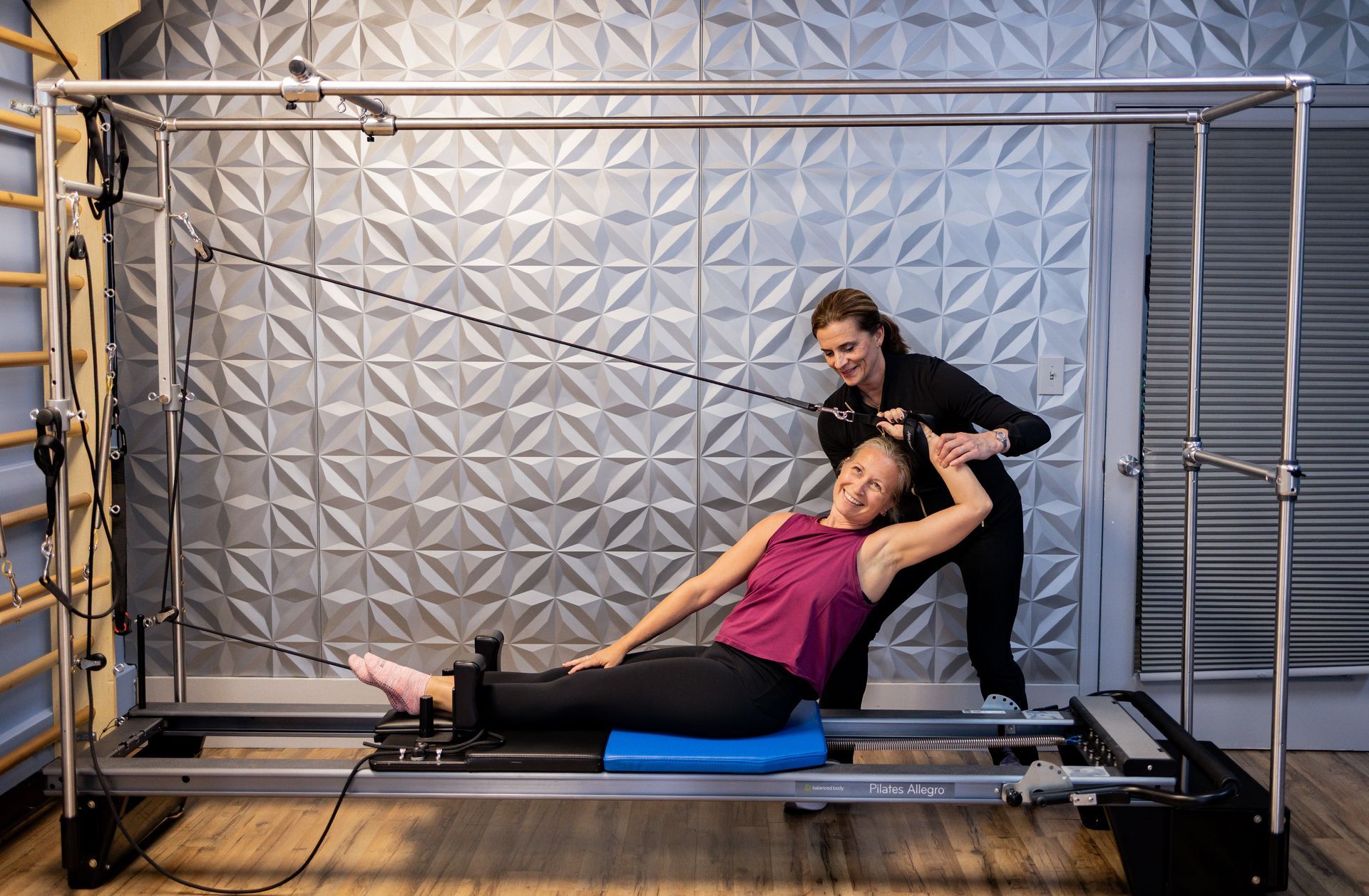 Woman assisting a person on a pilates reformer in a studio, with wall bars and exercise straps.