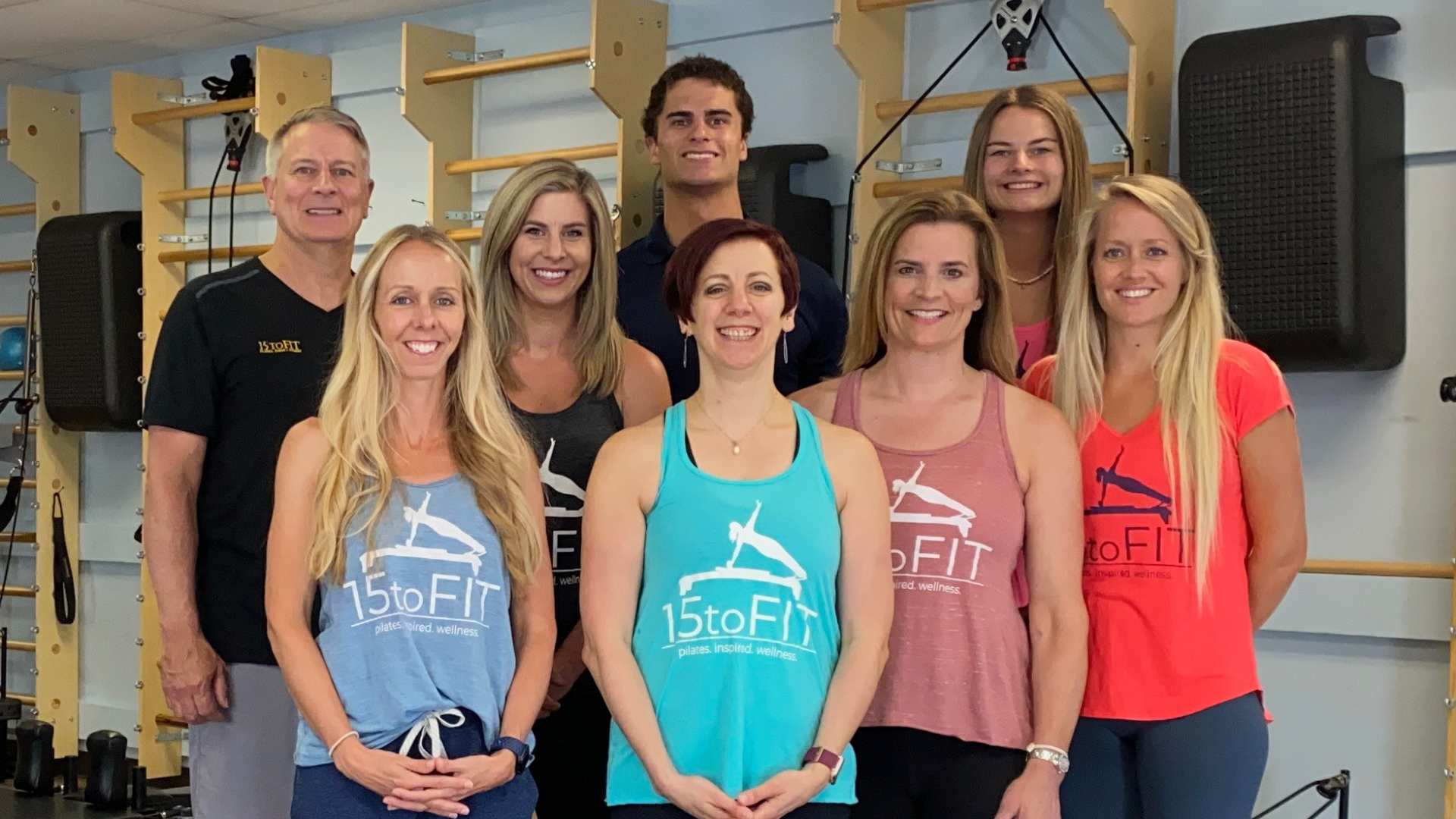 Seven people posing in a gym, wearing colorful workout shirts with a logo.
