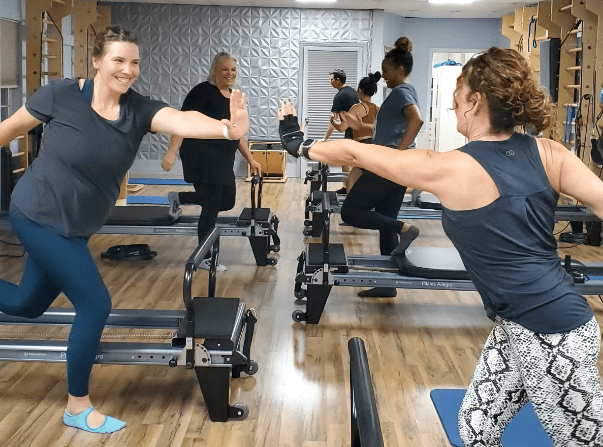 Woman using a resistance machine while lying on a reformer, guided by an instructor in a studio.
