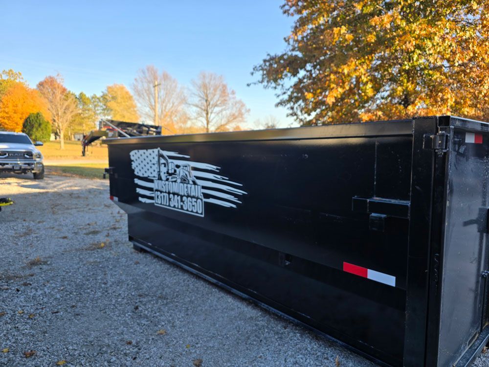 Black dump trailer with American flag graphic in yard with truck, trees, and blue sky.