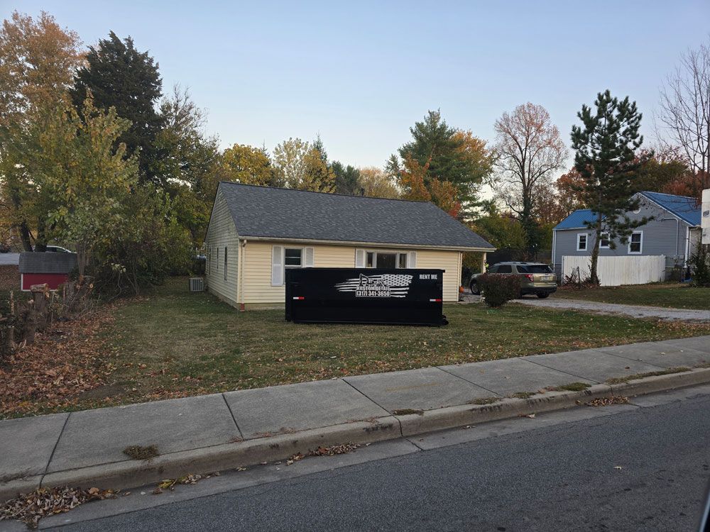 Yellow house with gray roof, black dumpster in front, trees in the background, blue sky.