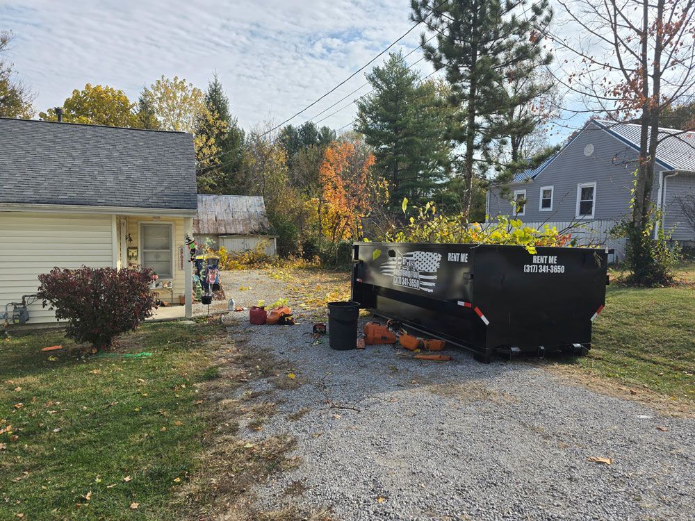 Black dumpster on gravel driveway near a house; yard work in progress.