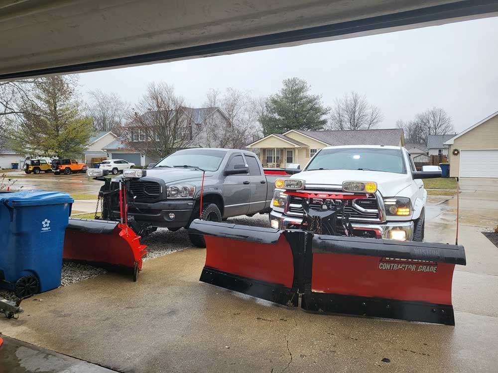 Two trucks with snow plows in a snowy driveway, facing forward.