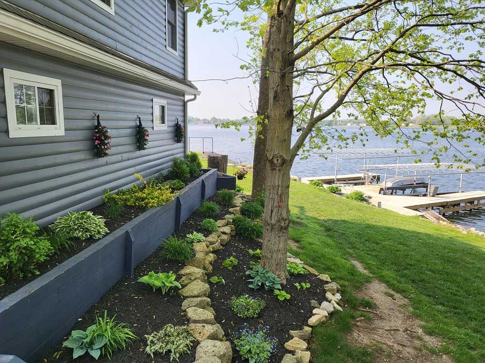 Blue house with a garden bed beside a body of water with docks and trees.